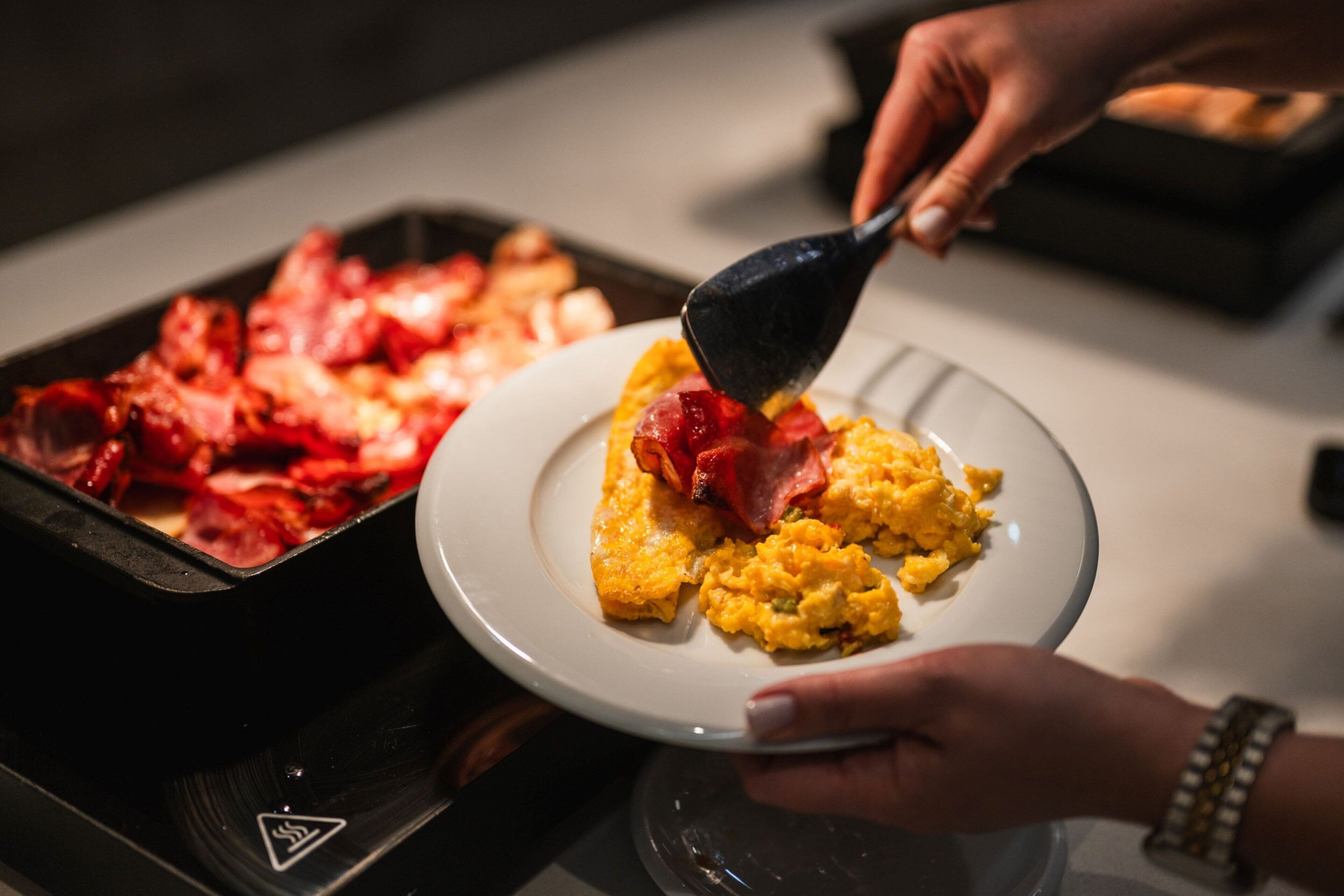 Una persona sirve huevos revueltos y tocino en un plato.