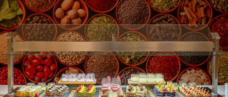 A dessert display with various sweets and pastries under a glass case, set against a backdrop of colorful bowls containing different ingredients.