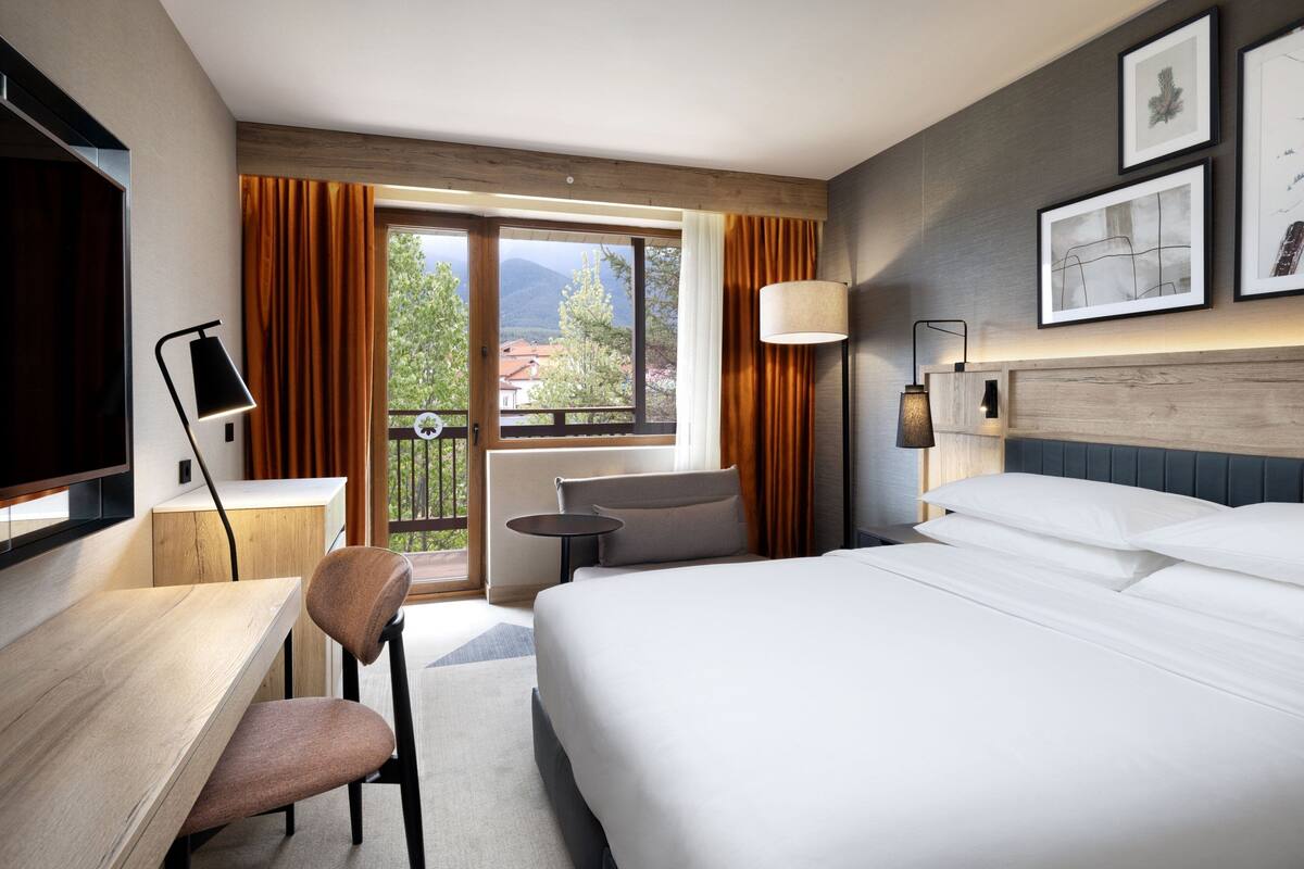 A hotel room with a bed, a desk, a chair, a small table, and a balcony with curtains.