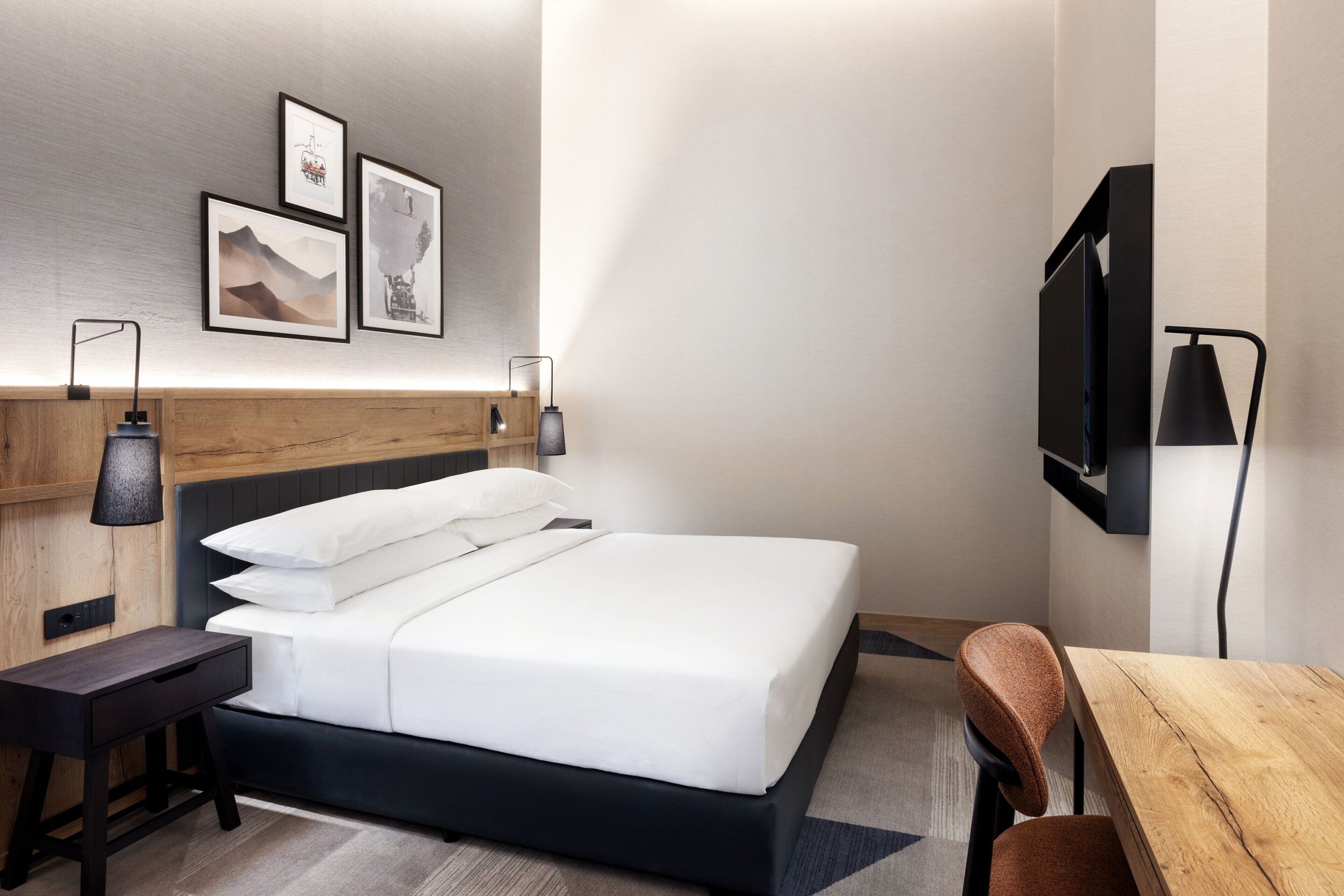 A modern hotel room with a wooden headboard, a large bed with white linens, a desk with a lamp, a TV mounted on the wall, and framed artwork above the bed.