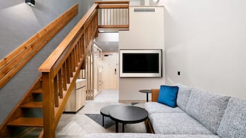 A modern living room with a wooden staircase, a grey sofa, a small round table, and a flat-screen TV mounted on the wall.