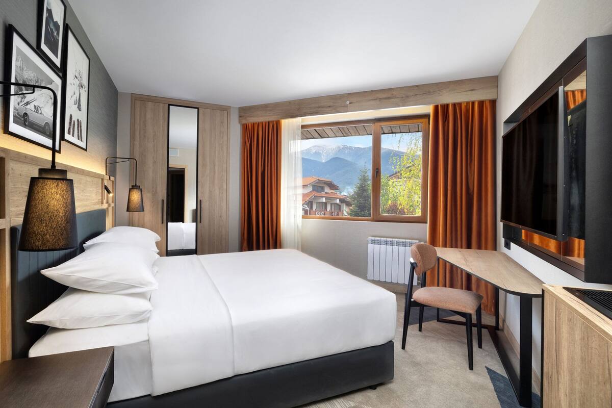 A hotel room with a bed, a desk, a chair, a TV, and a window with a mountain view.