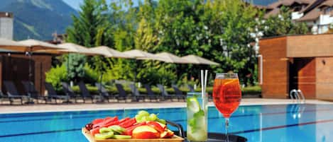 A poolside table with a fruit platter and drinks, surrounded by lounge chairs and a mountain view.
