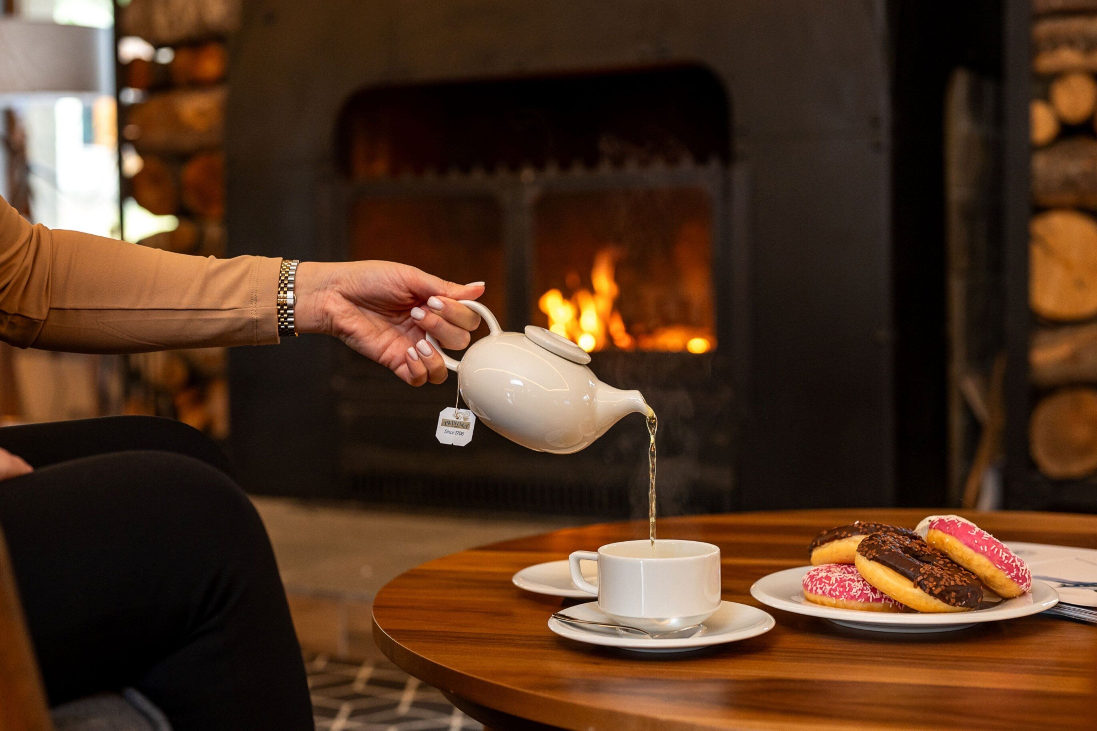 A hand pouring tea into a cup on a table with donuts.