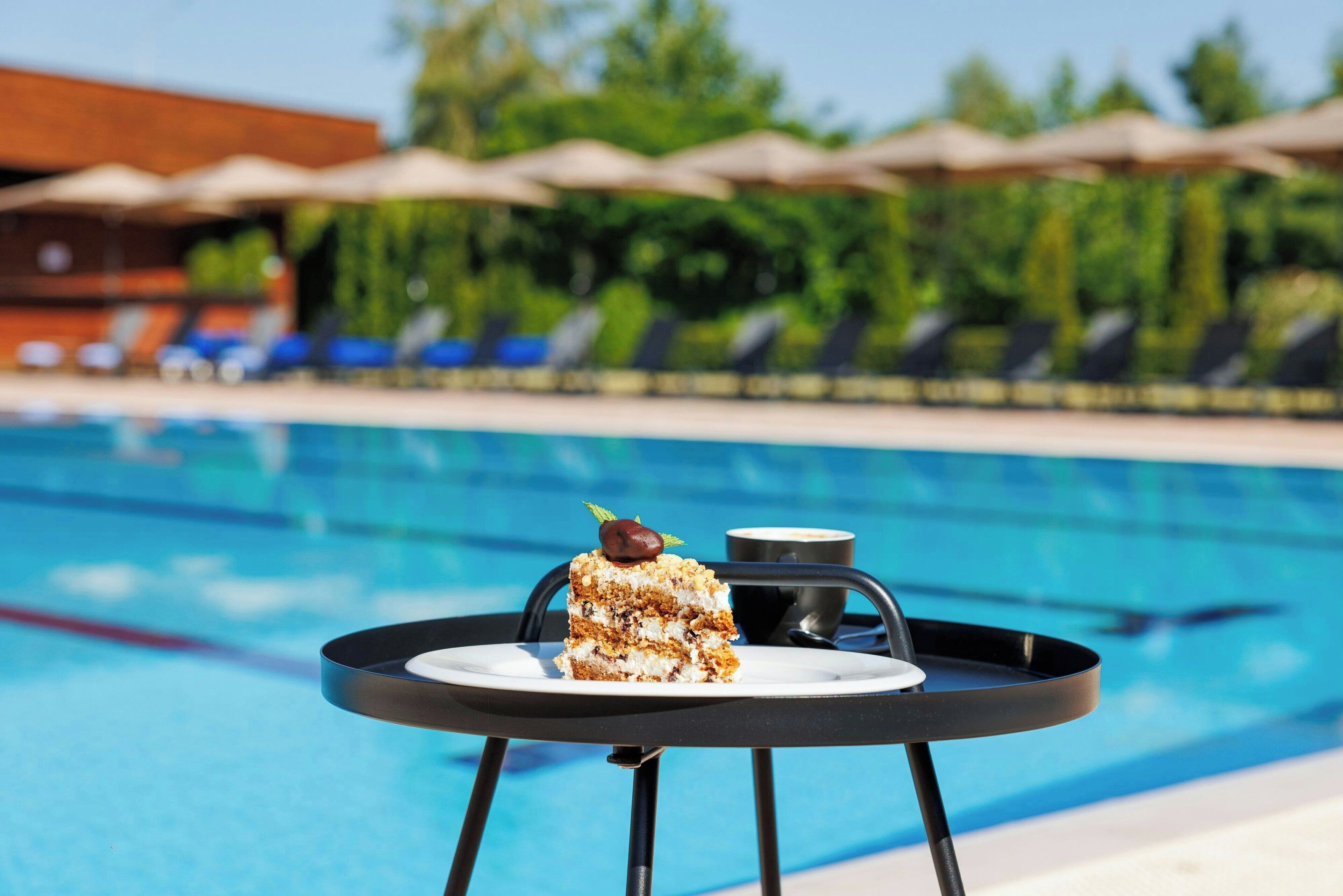 A slice of cake on a tray by a poolside table with a drink, surrounded by lounge chairs and umbrellas.