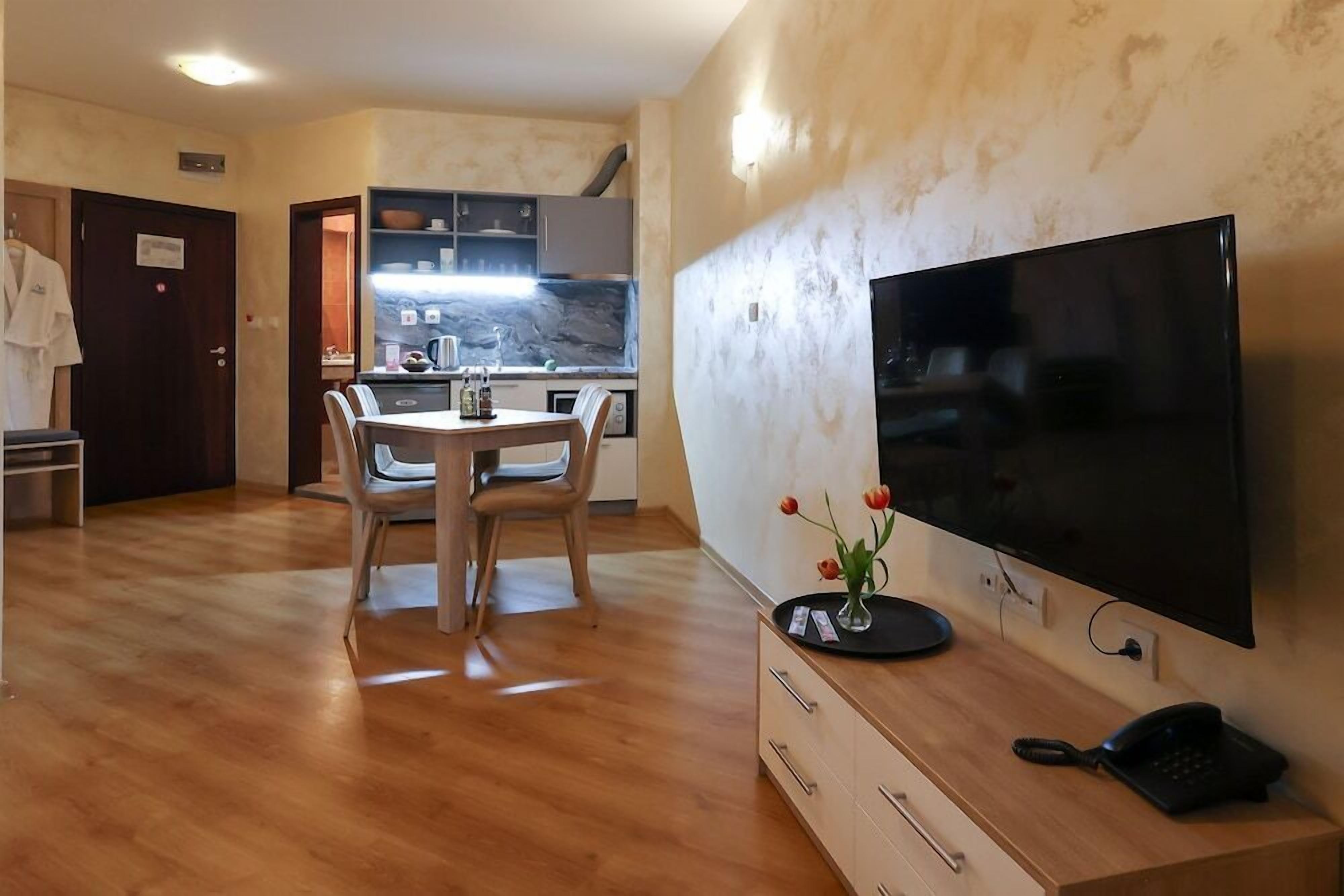 Un soggiorno moderno con zona pranzo, una TV a schermo piatto montata a parete e una cucina visibile attraverso una porta aperta.