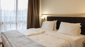 Ett hotellrum med en stor säng, vita sängkläder, en sänggavel i trä, en sänglampa och utsikt över staden genom tunna, genomskinliga gardiner.