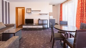 Una habitación de hotel con sofá, una mesa de comedor con sillas, un televisor, un mueble bajo y una ventana con cortinas.