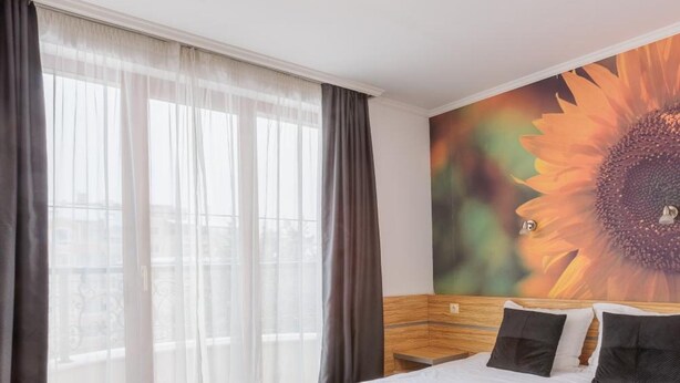 Habitación de hotel con una cama grande, cabecera de madera, ventana con cortinas translúcidas y un mural de girasol en la pared.
