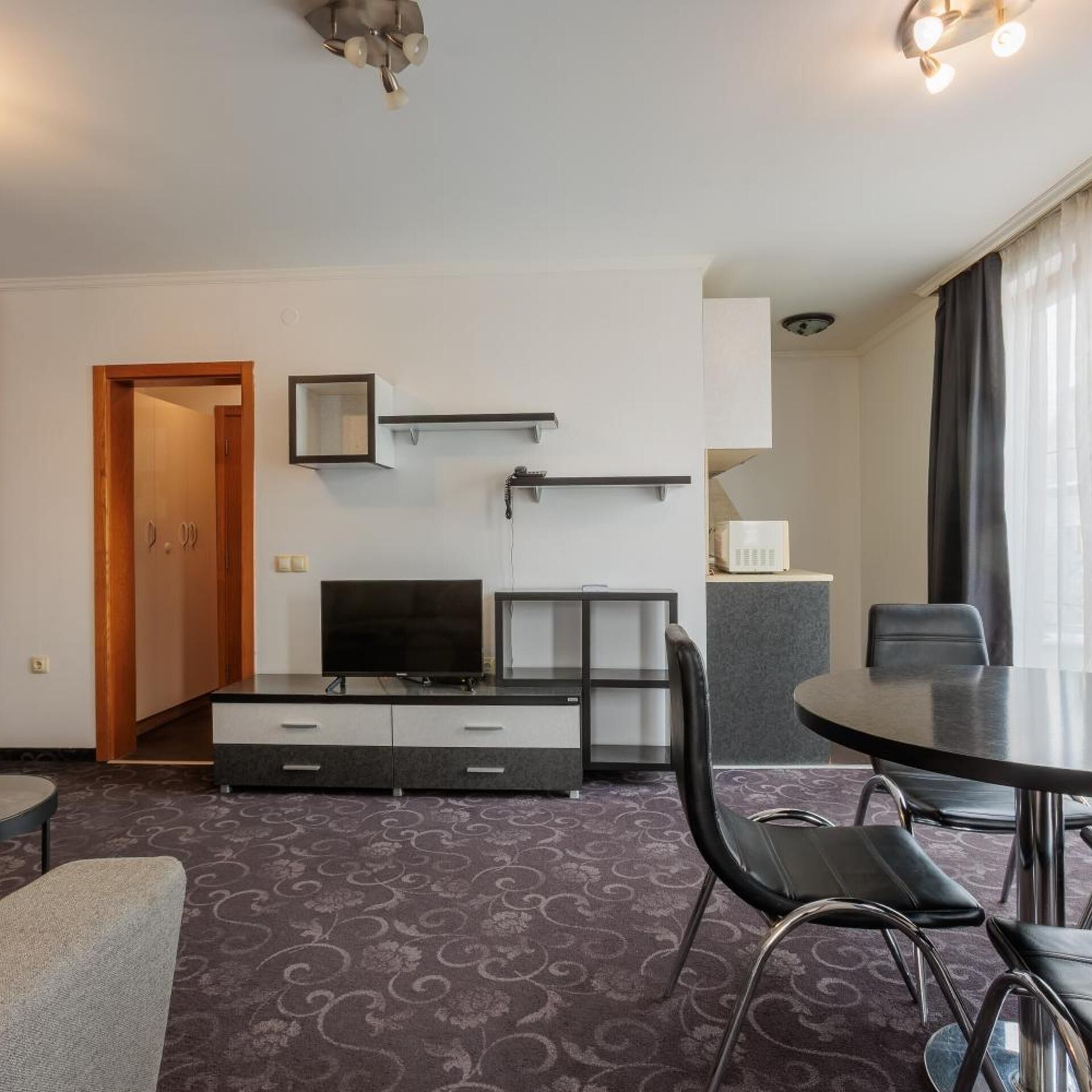 A hotel room with a flat-screen TV, a desk with a chair, a dining table with chairs, a mirror, and a wardrobe.