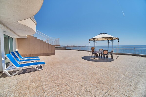 Ett soligt utomhusområde med en pool, solstolar, en pergola och utsikt över havet.