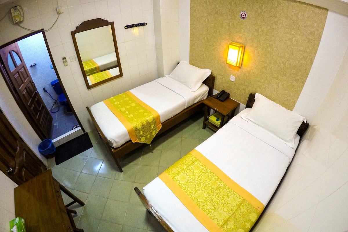 A hotel room with two single beds, a wooden nightstand with a phone and a small table, a mirror, and a wall-mounted light fixture.