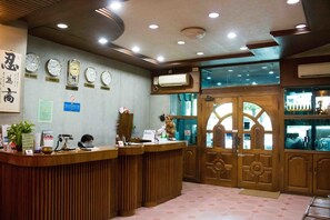 A reception area with a wooden reception desk, multiple clocks displaying time from different cities, an air conditioning unit, and a glass door leading to the outside. - May Shan Hotel (Yangon)