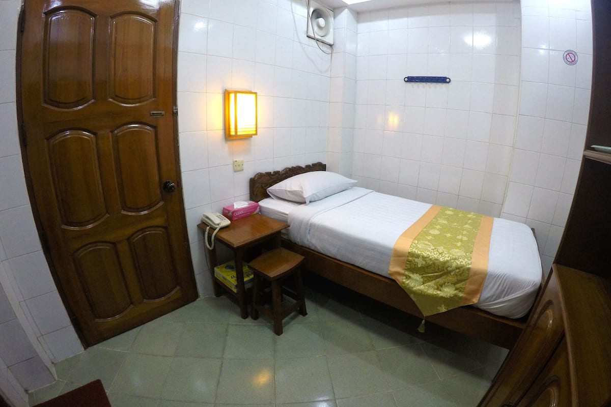 A small, clean room with a single bed, a wooden desk, a chair, a bedside table, and a wall-mounted light.