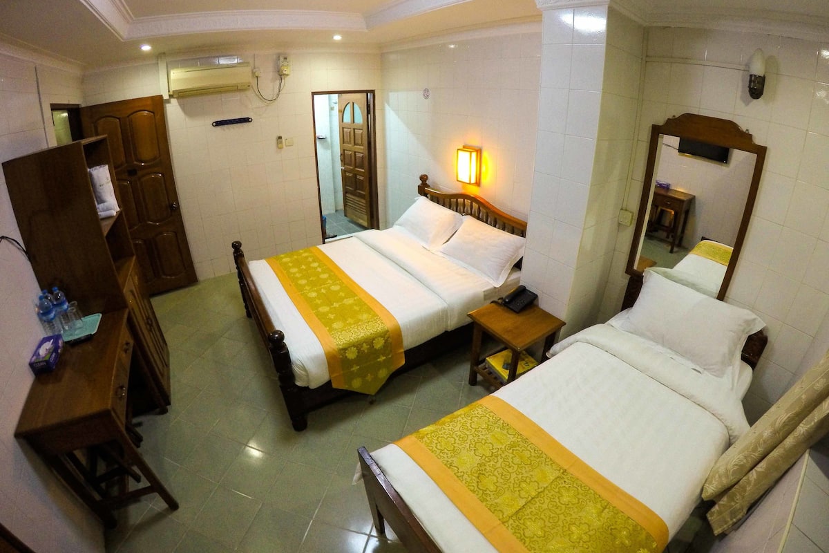 A hotel room with two beds, a wooden wardrobe, a bedside table with a lamp, and a mirror on the wall.