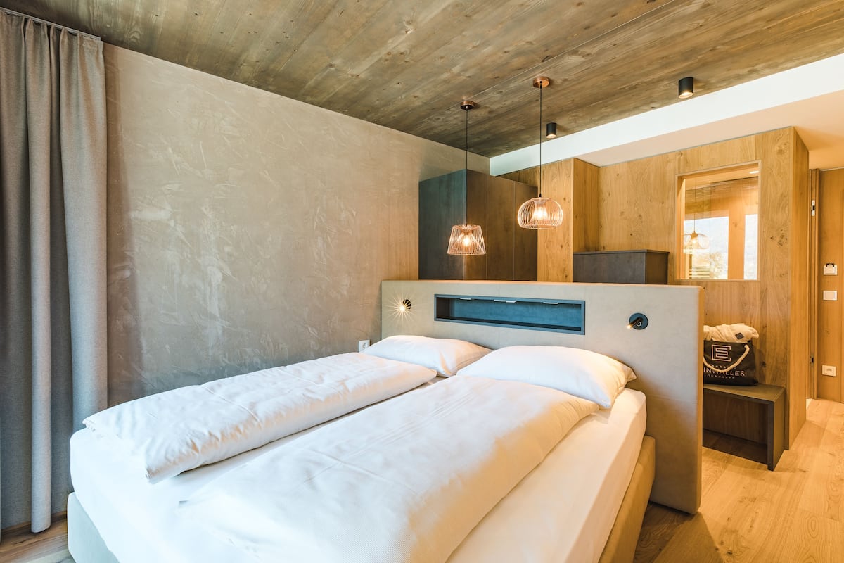 Un dormitorio moderno con techo de madera, una cama con ropa blanca, una mesita de noche y una lámpara colgante.
