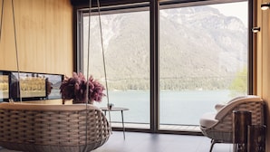 Ein modernes Zimmer mit großem Fenster mit Blick auf einen See und Berge, einem Korbsessel, einem kleinen Tisch mit Tasse und einem Hängesessel.