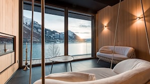 Ein modernes Zimmer mit großem Fenster mit Blick auf einen See und Berge, einem Hängesessel, einem kleinen runden Tisch und einem Sofa.