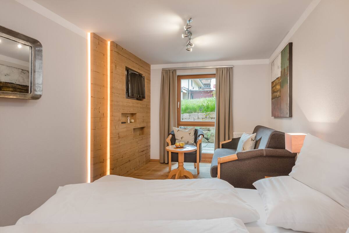 Une chambre d’hôtel comprenant un lit, une petite table avec une chaise, un canapé, un téléviseur fixé au mur et une fenêtre avec des rideaux.
