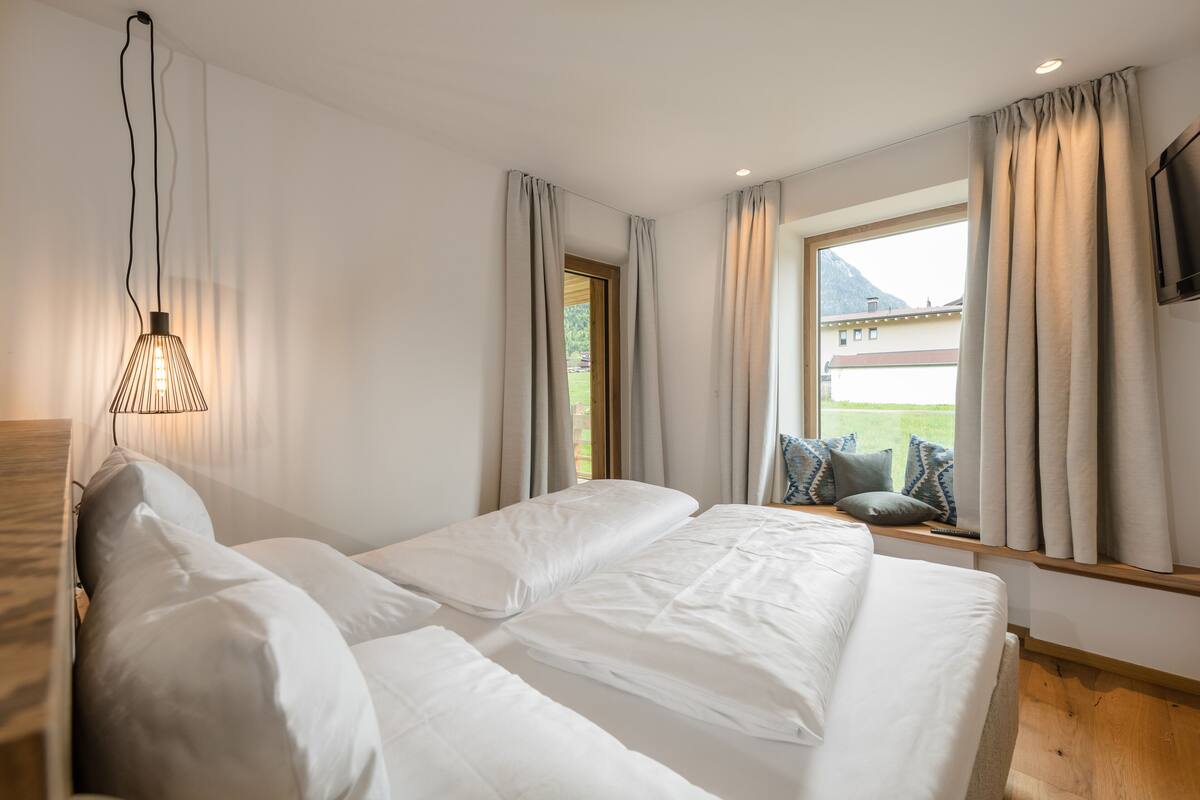 Une chambre avec un grand lit, une fenêtre avec des rideaux, un téléviseur fixé au mur et un luminaire suspendu.