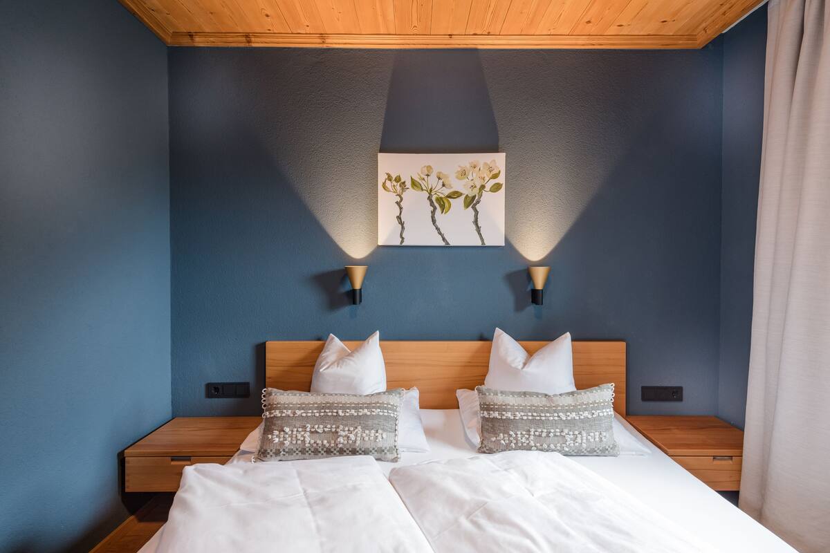 Une chambre d’hôtel avec une tête de lit en bois, deux tables de chevet, un lit avec du linge de lit blanc, des coussins décoratifs et une œuvre d’art représentant des fleurs accrochée au mur.