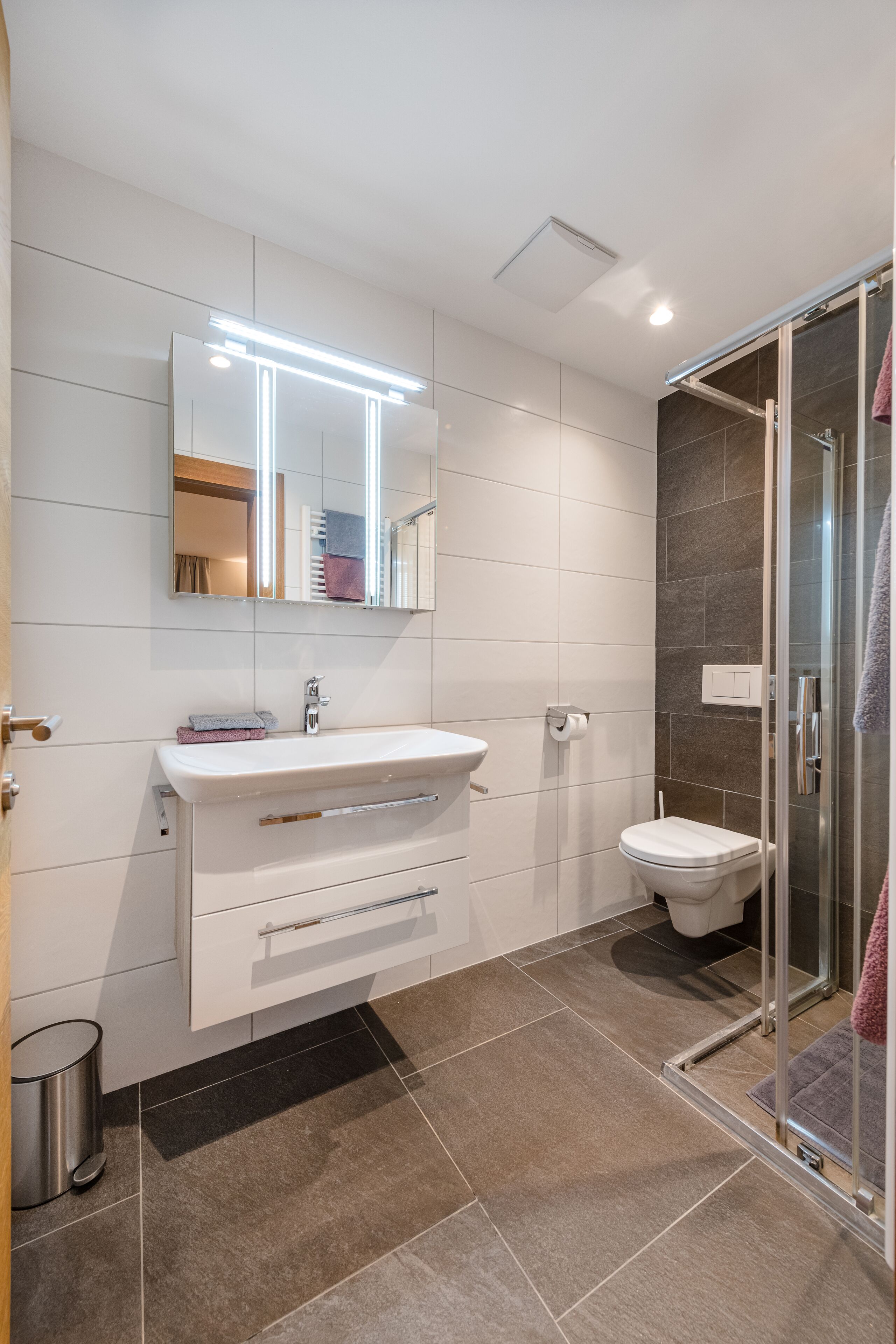 A modern bathroom with a white sink, mirror, and toilet. It includes a glass shower enclosure and a black mat on the floor.