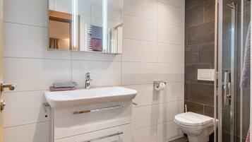 Un baño moderno con un lavamanos blanco, espejo e inodoro. Cuenta con un box de ducha de vidrio y una alfombra negra en el suelo.