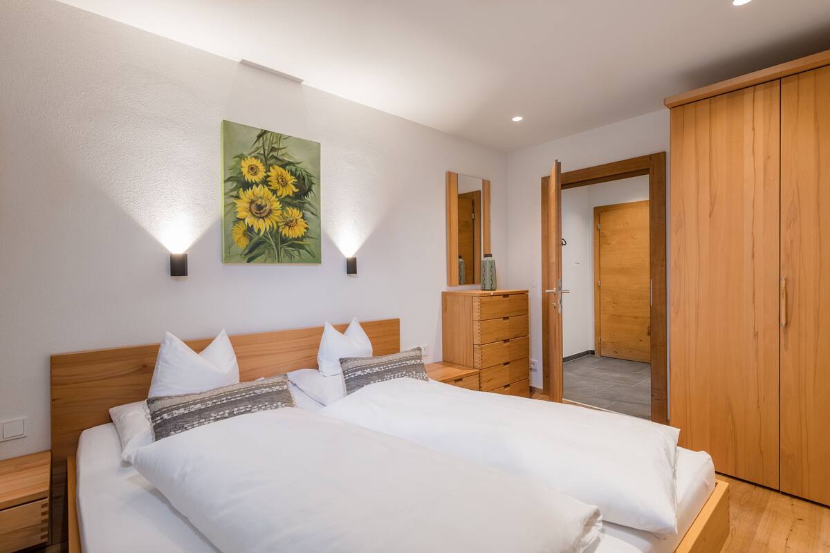 Une chambre à coucher avec un lit double, une tête de lit en bois, des tables de chevet, une commode, un miroir et une porte donnant sur une autre pièce.