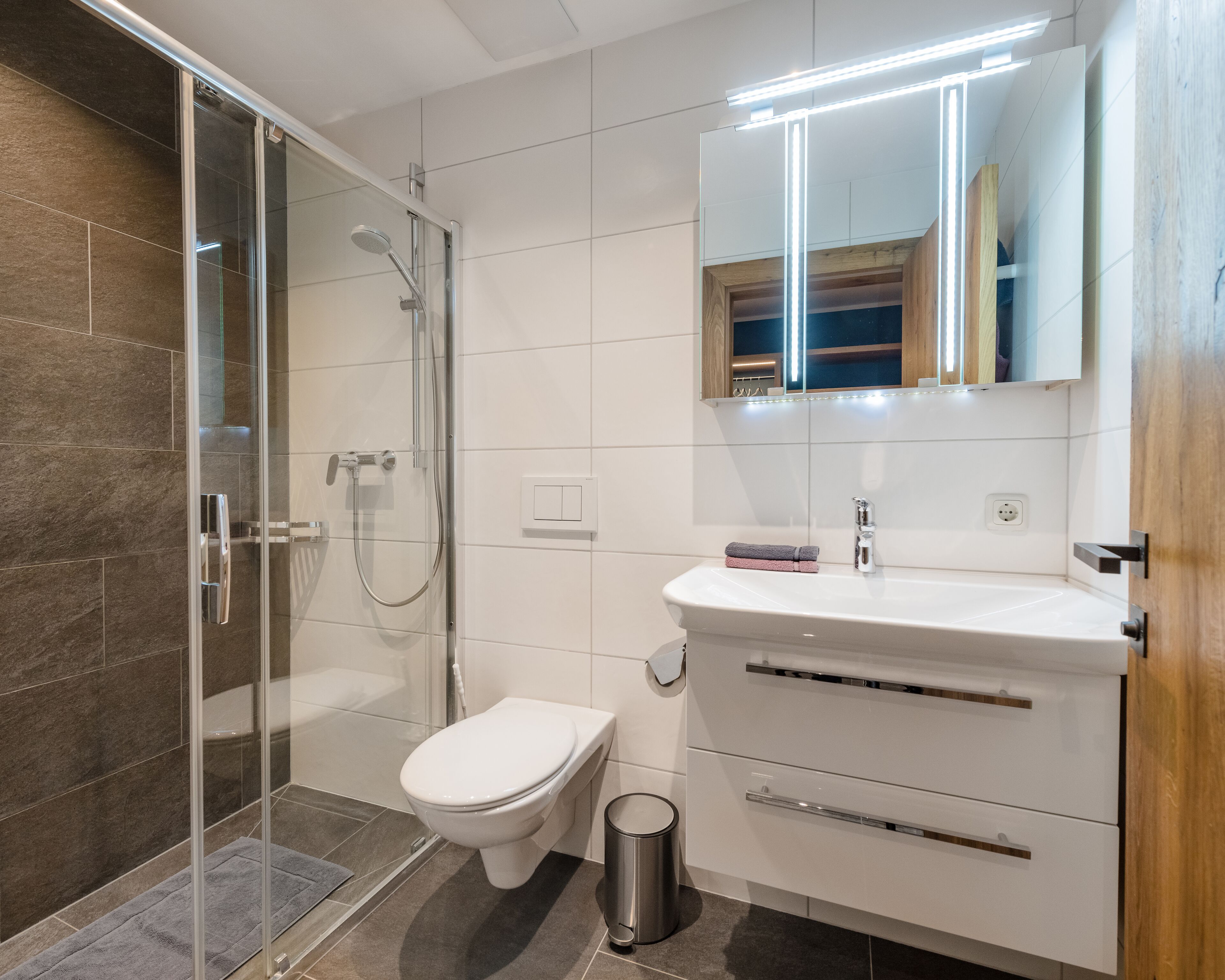 Un baño moderno con cabina de ducha de vidrio, inodoro blanco, tocador con espejo y un cubo de basura.