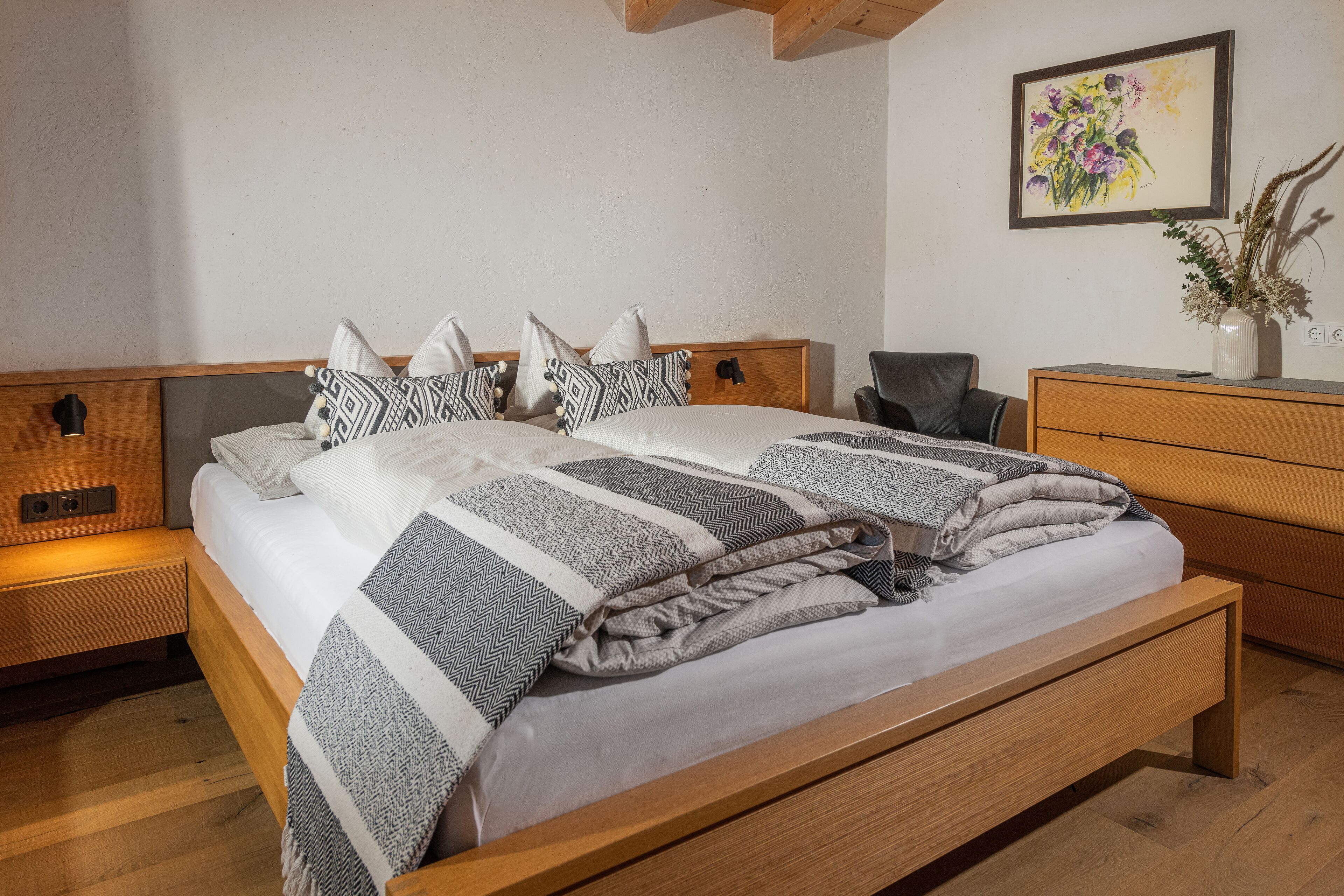 Una cama bien hecha con ropa de cama estampada, una mesita de noche con lámpara, un aparador de madera y un cuadro colgado en la pared.