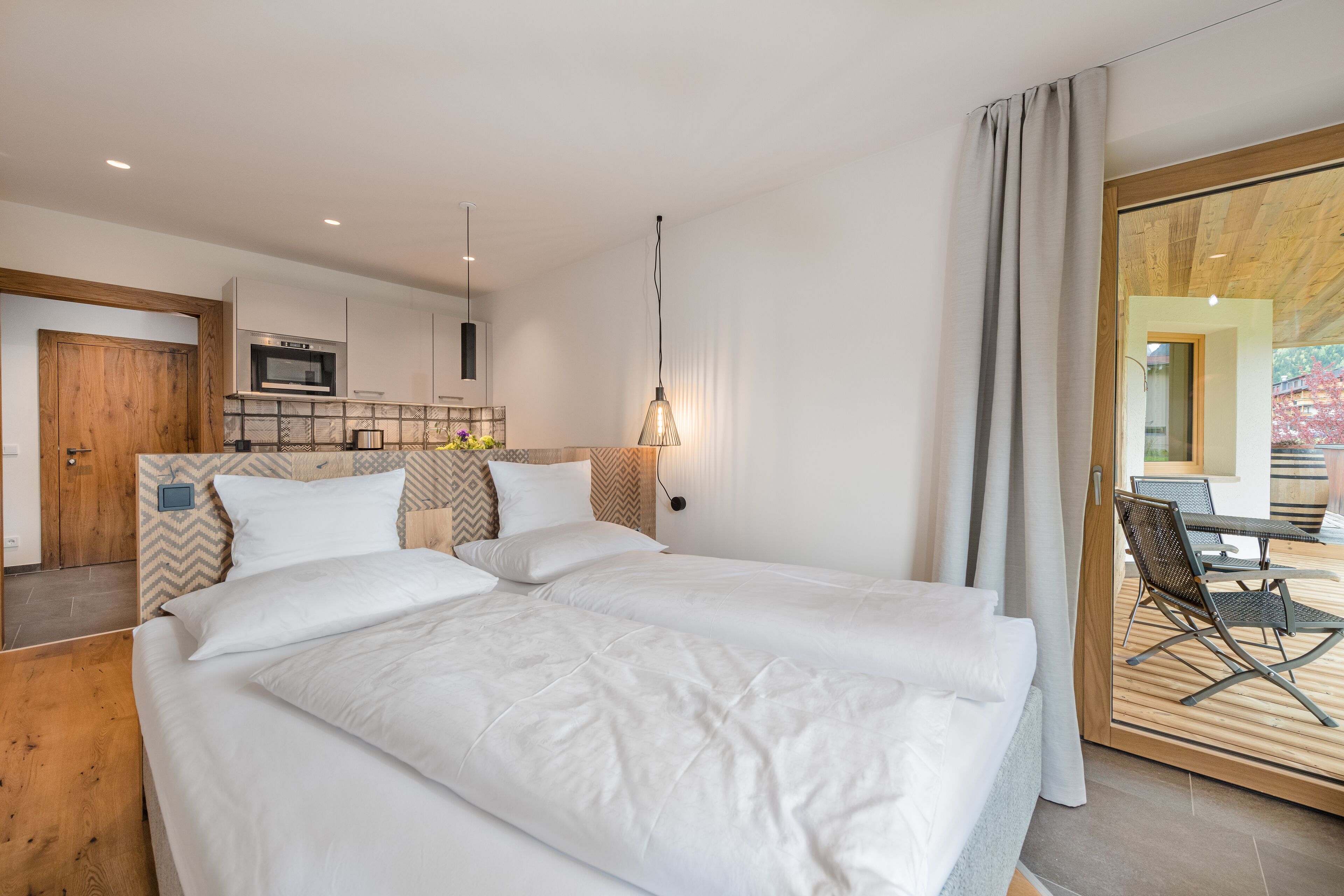 Una habitación de hotel moderna con una cama grande, cabecera de madera, zona de cocina y balcón con mobiliario exterior.