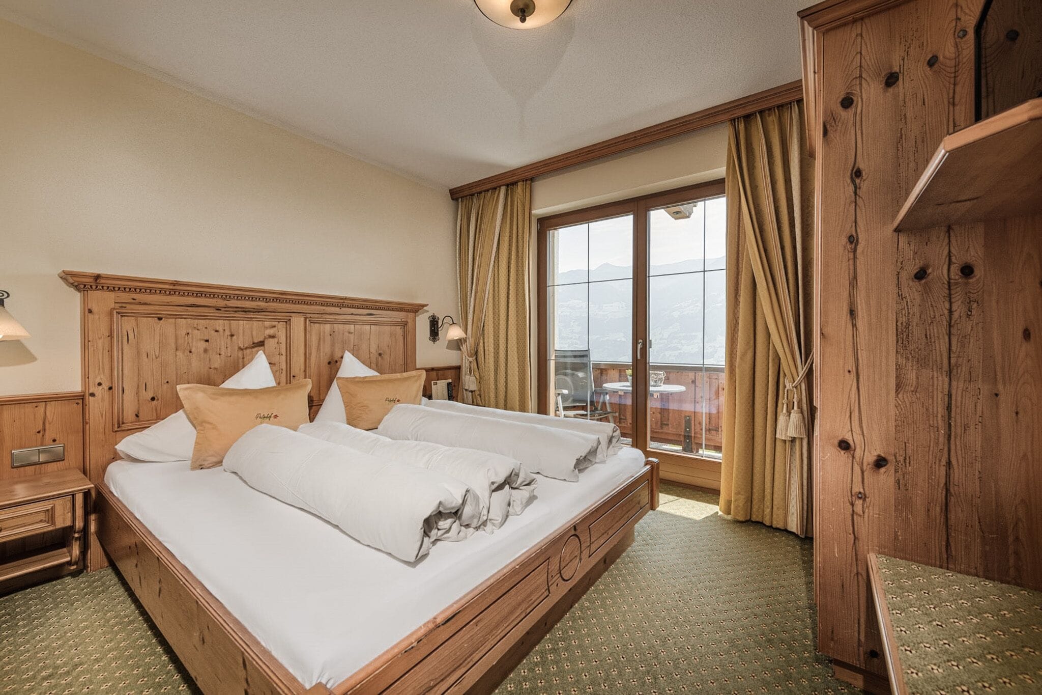 Una cama doble con cabecera de madera, una mesita de noche, una lámpara y vistas a un balcón con cortinas.