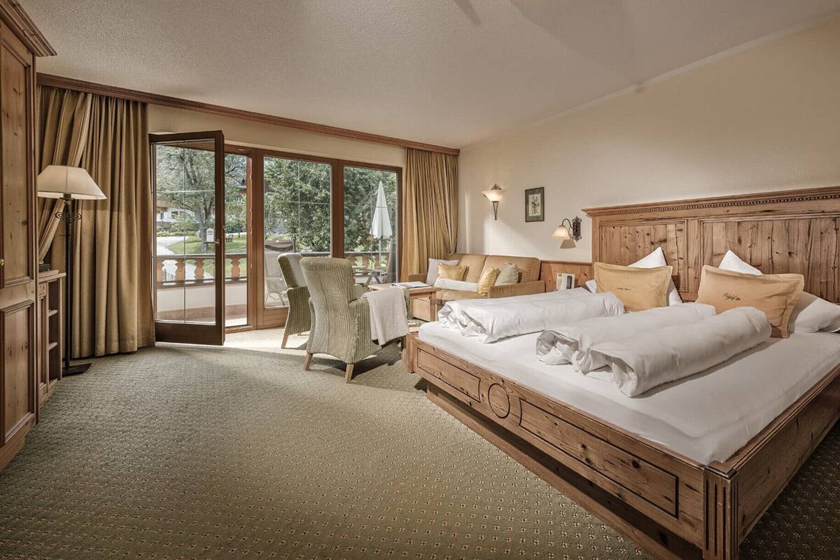 Une chambre d’hôtel spacieuse, dotée d’un grand lit, d’un coin salon avec un fauteuil et une table, et offrant une vue sur l’extérieur grâce à une porte vitrée.