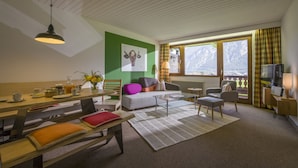A cozy living room with a wooden bench, a sofa, a coffee table, and a TV. There is a window with a view of mountains and a lamp on the side table.
