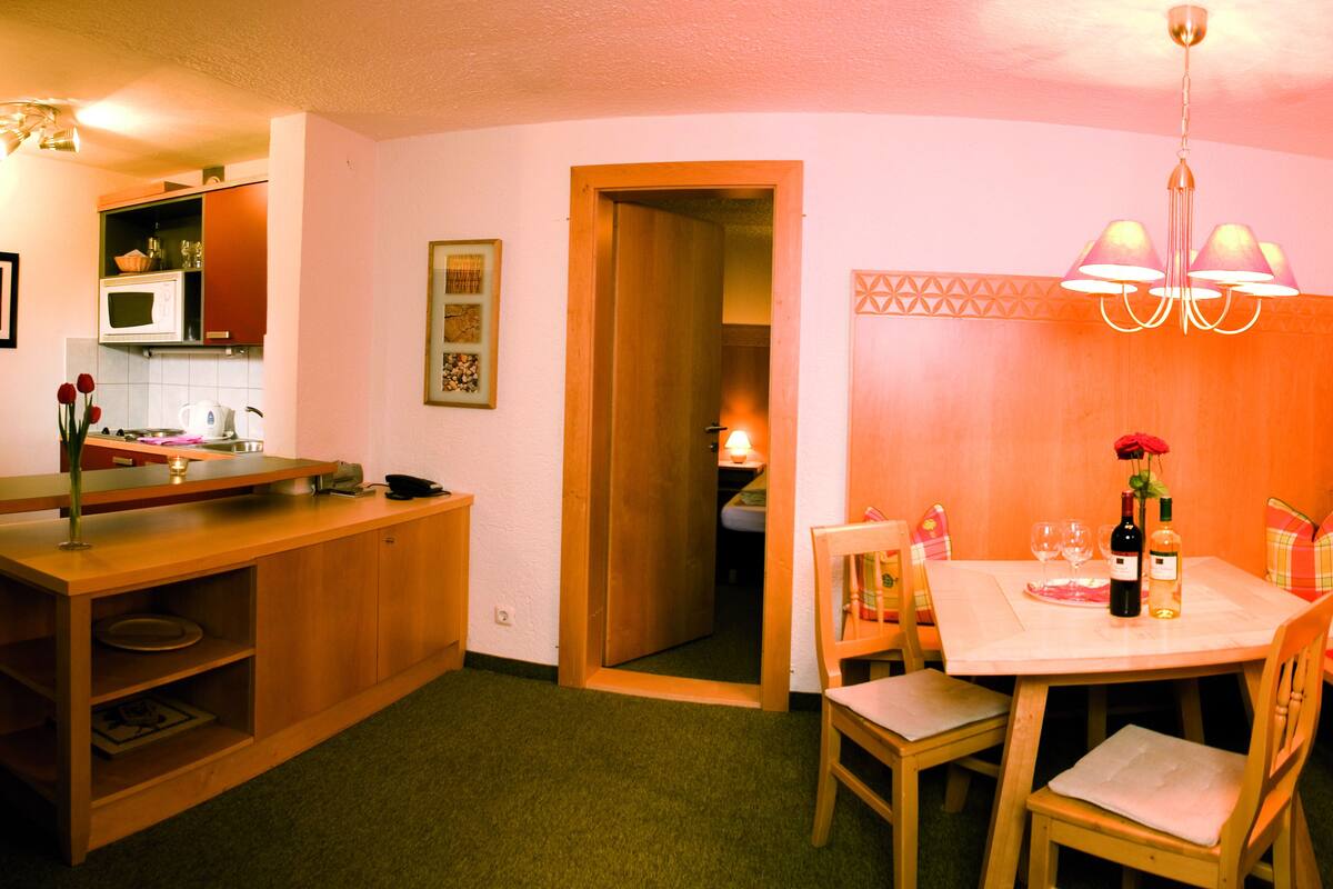 A compact kitchen with a microwave, sink, and wooden cabinets. There is a dining area with a table, chairs, and a bench.