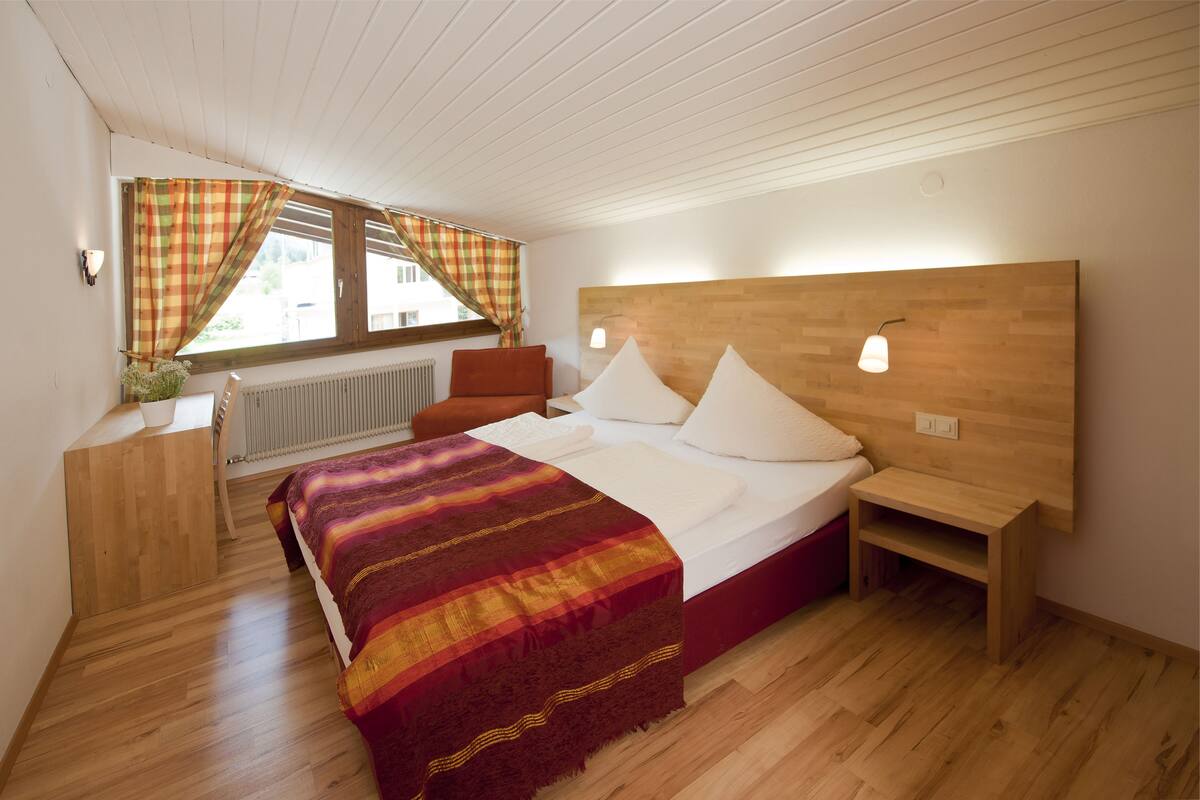 A bedroom with a wooden headboard, a bed with white bedding, a red and yellow striped blanket, a wooden nightstand, and a window with curtains.