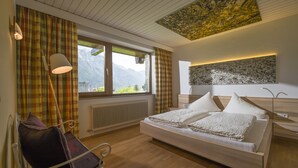 A bedroom with a bed, a chair, a radiator, a window with curtains, and a view of mountains.