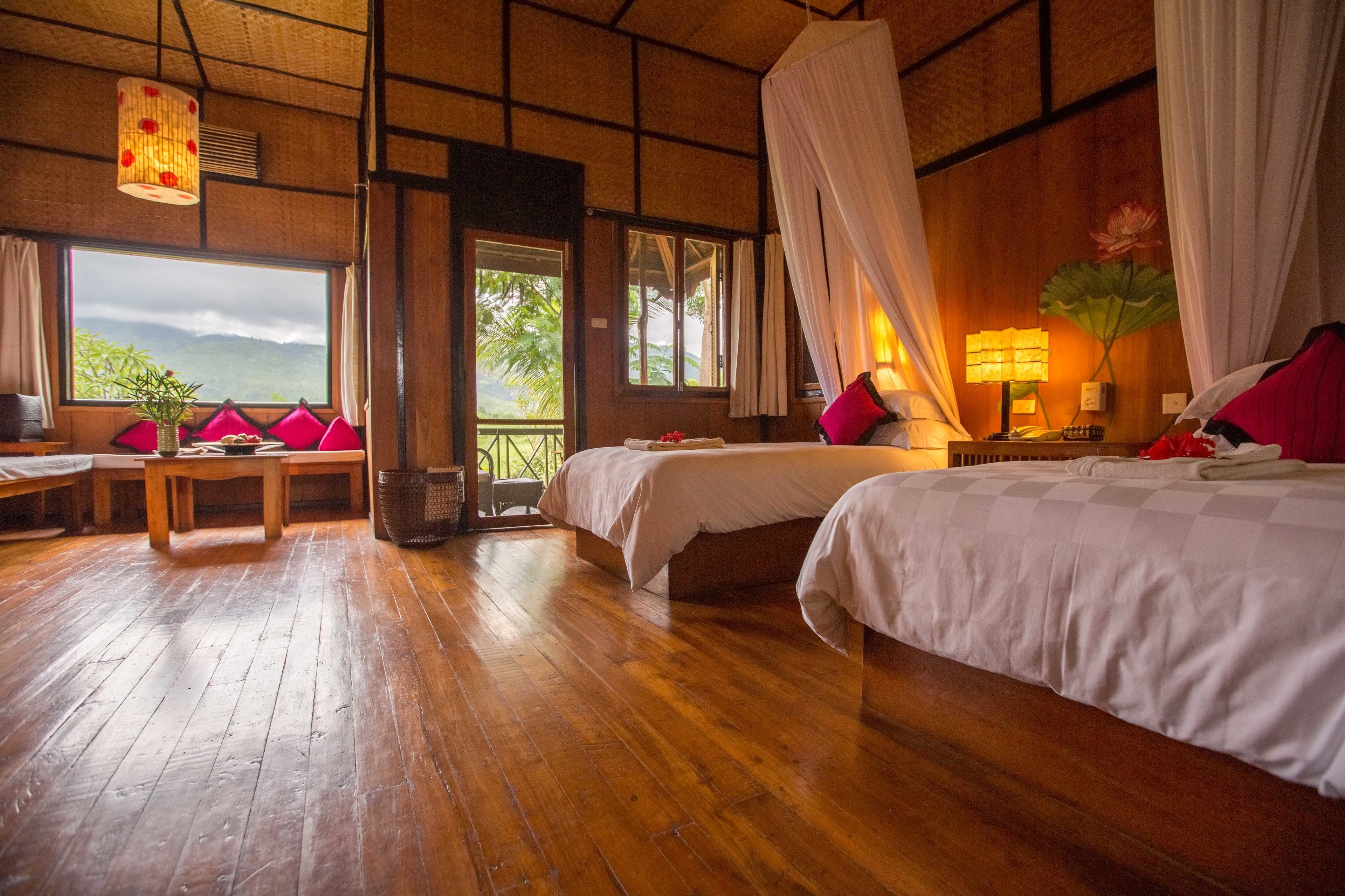 A room with two beds, a wooden floor, a table with chairs, and a view of the outdoors.