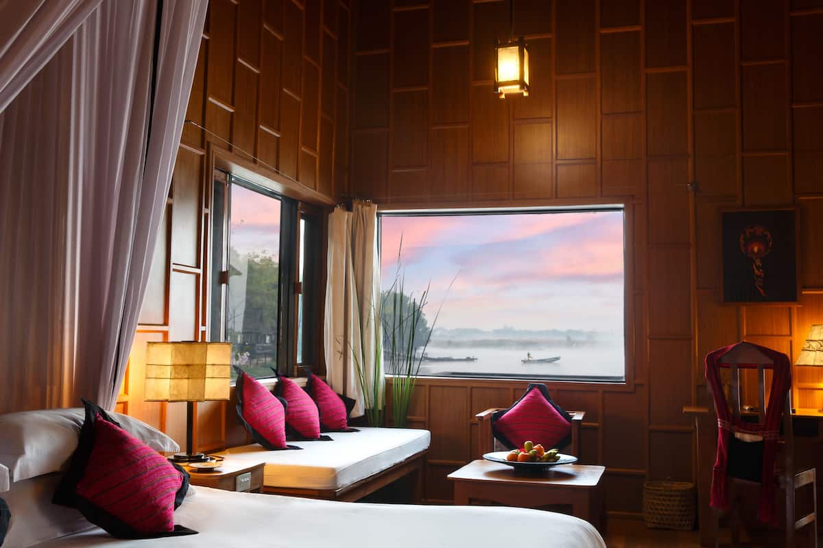 A hotel room with a bed, a small table with a bowl of fruit, a chair, a window with a view of a body of water, and a lamp on the nightstand.
