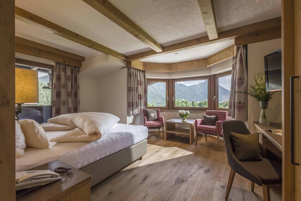 A bedroom with a large bed, a desk, a chair, and a view of mountains through the windows.