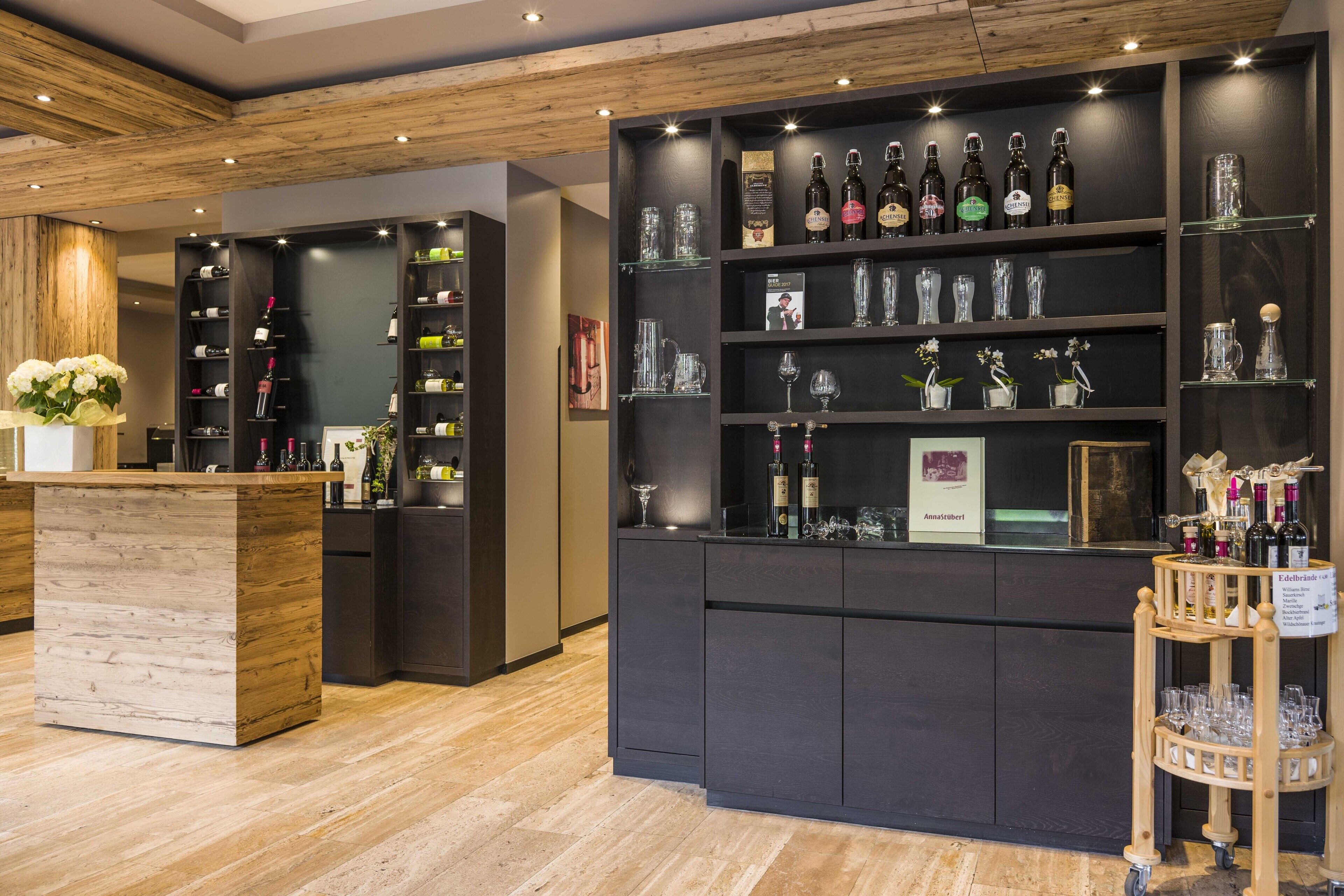 Una zona de bar con estantes llenos de botellas y vasos, una barra de madera y un pequeño carrito con copas.