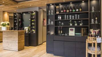 Una zona de bar con estantes llenos de botellas y vasos, una barra de madera y un pequeño carrito con copas.