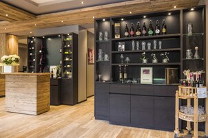Una zona de bar con estantes llenos de botellas y vasos, una barra de madera y un pequeño carrito con copas.