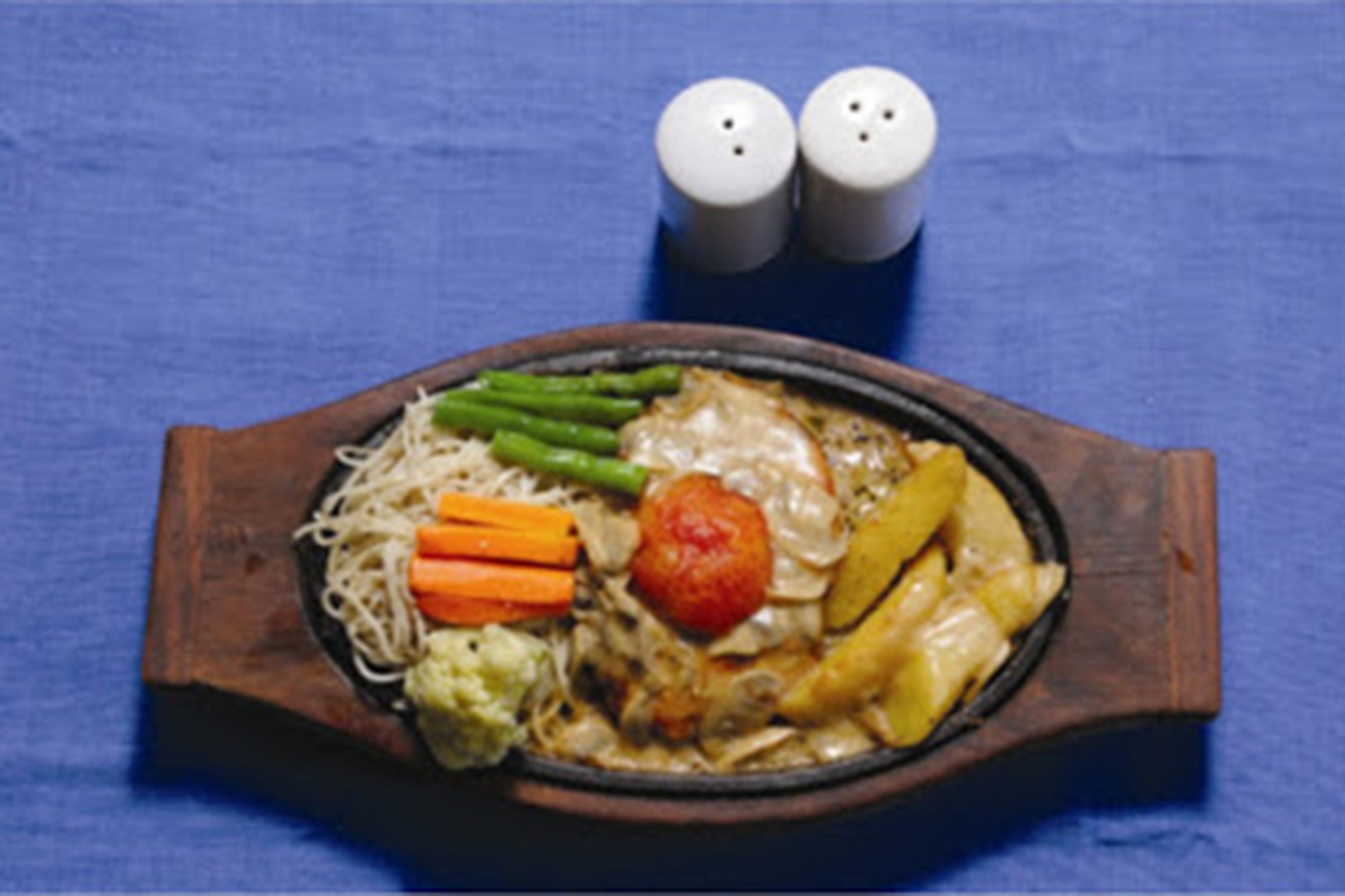Une assiette de nourriture comprenant des nouilles, des légumes et un morceau de viande, le tout présenté sur une planche en bois.