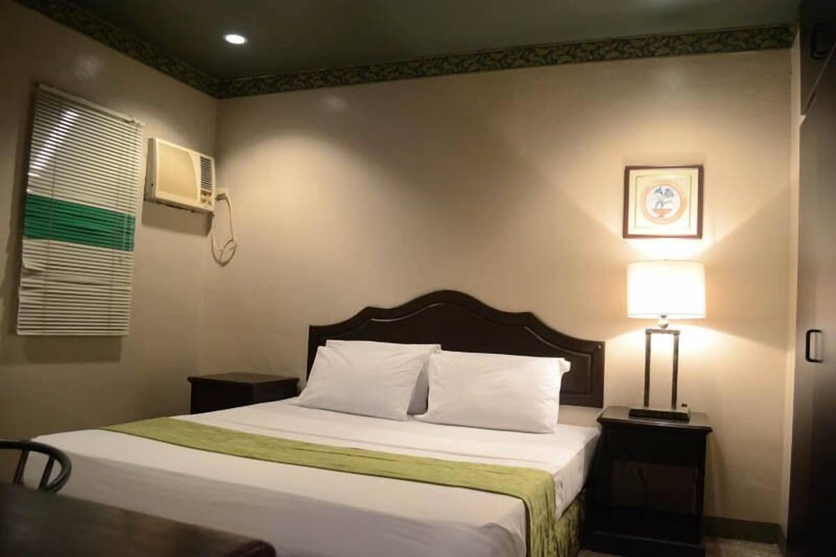 A hotel room with a bed, bedside tables, a lamp, a framed picture on the wall, and an air conditioning unit.