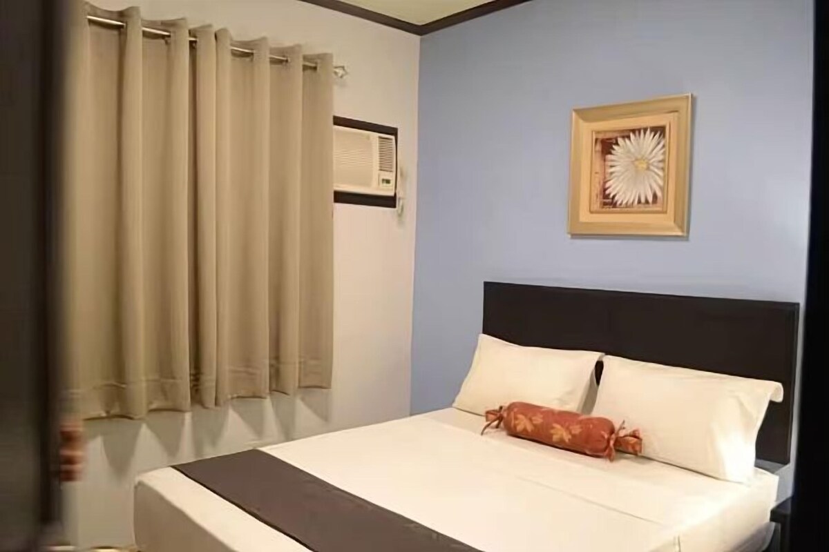 A bedroom with a bed, beige curtains, a wall-mounted air conditioner, and a framed flower decoration.