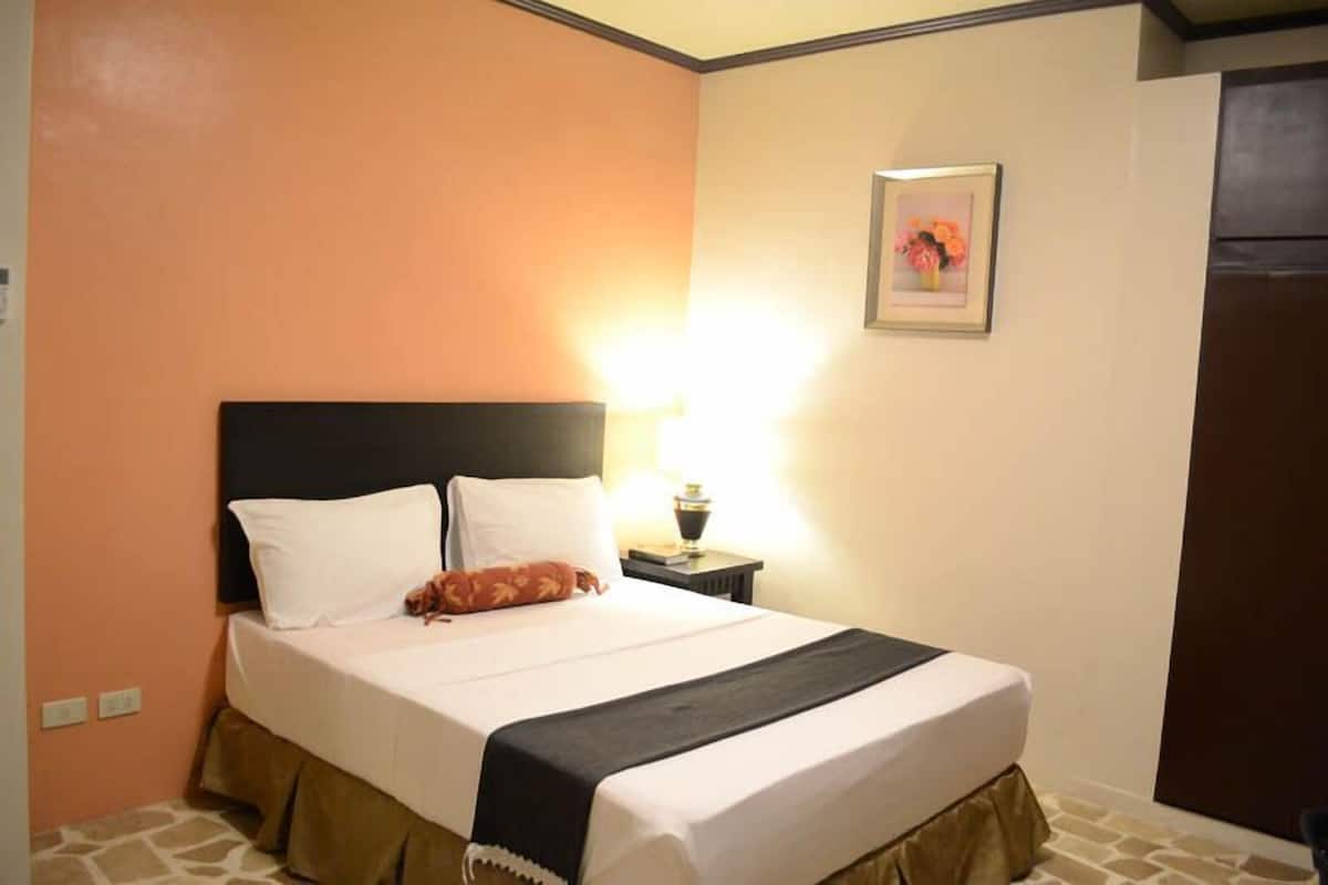 A neatly made bed with white linens, a black headboard, and a brown skirt. There is a small bedside table with a lamp and a framed picture on the wall.