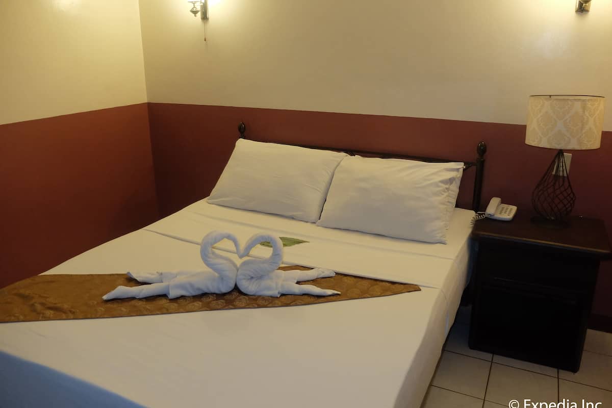 A hotel room with a neatly made bed, a heart-shaped towel arrangement, a bedside lamp, and a phone on the nightstand.
