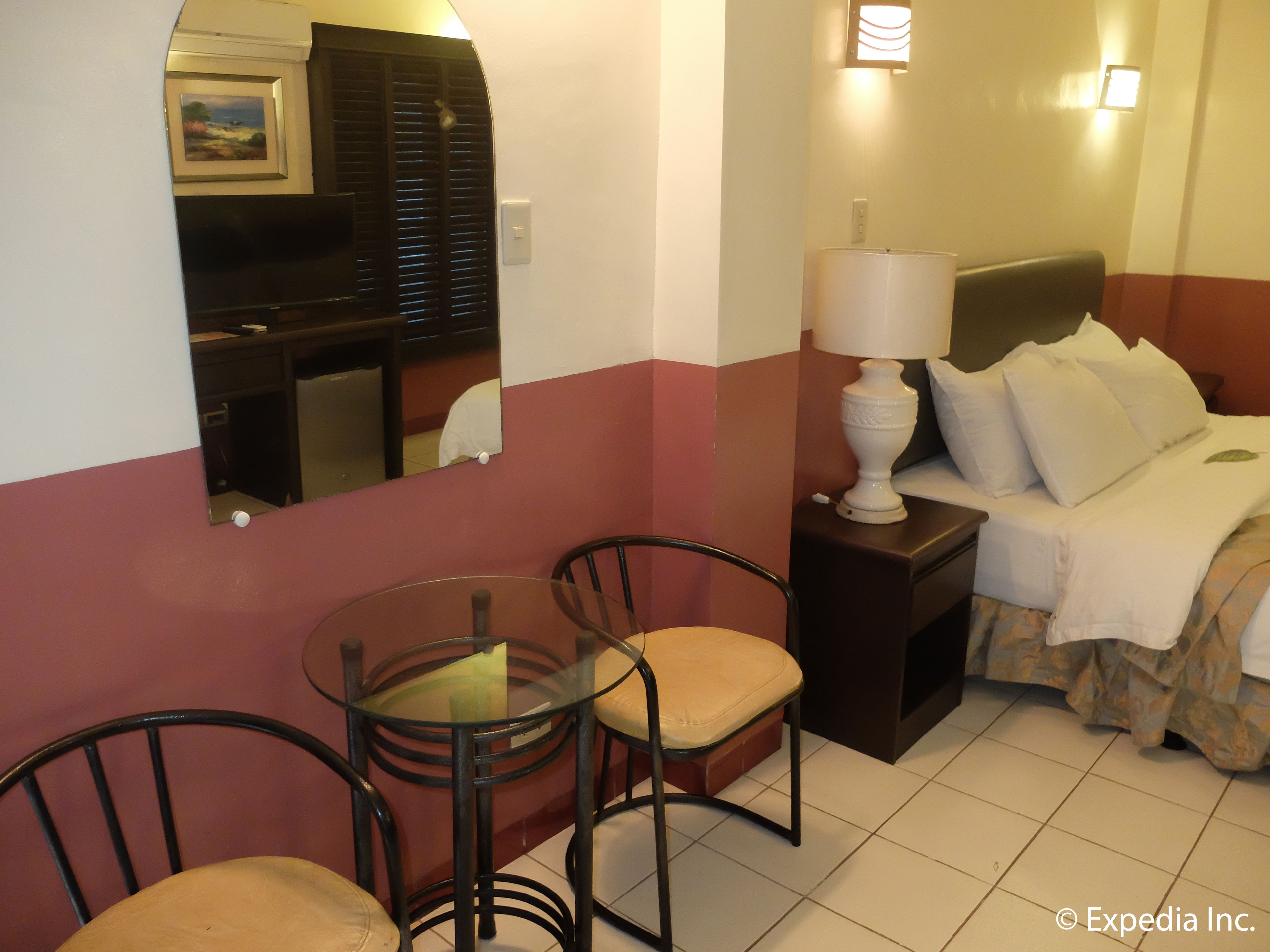 A hotel room with a bed, bedside lamp, two chairs, a small table, and a mirror reflecting a hallway.