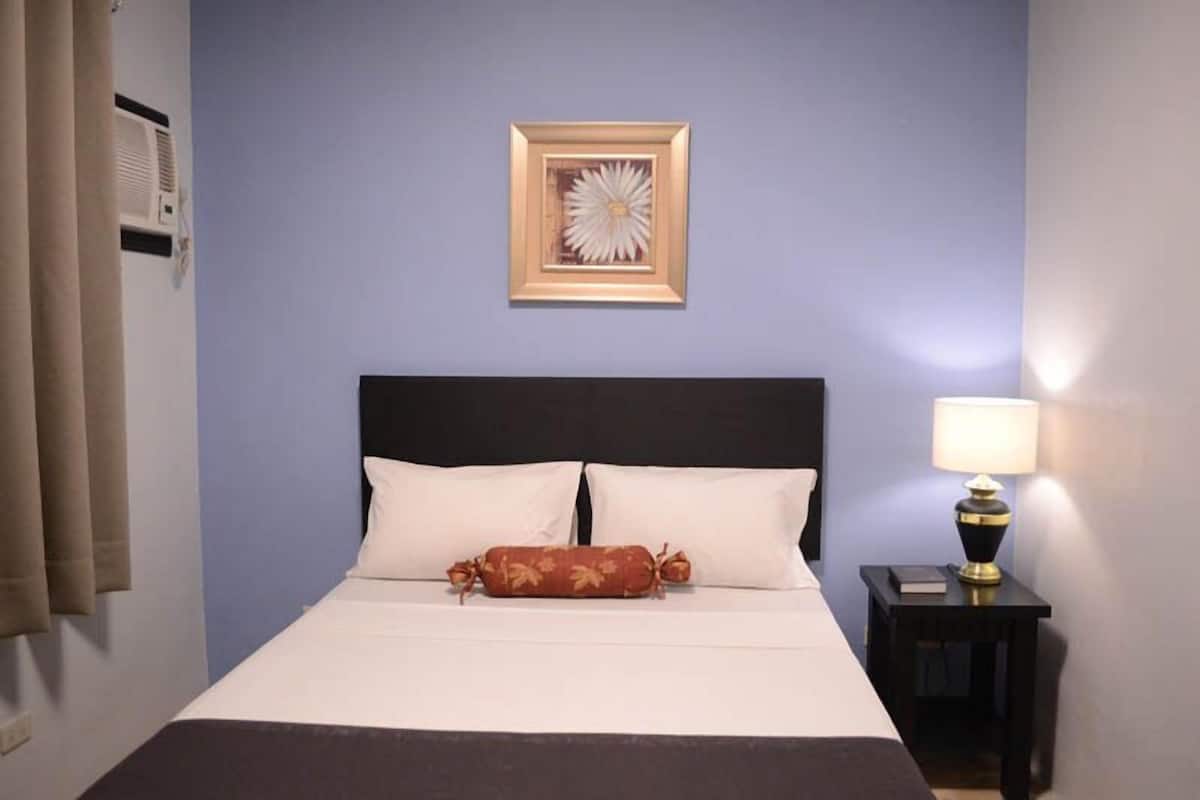A hotel room with a bed, a framed picture on the wall, a bedside lamp, and a window with curtains.